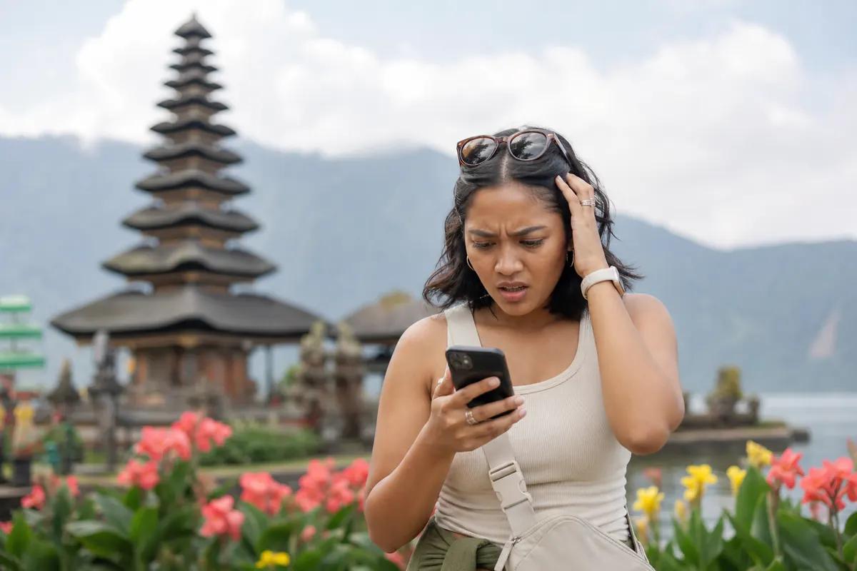 Stunned traveler staring at her smartphone with a Balinese temple and flower beds behind her