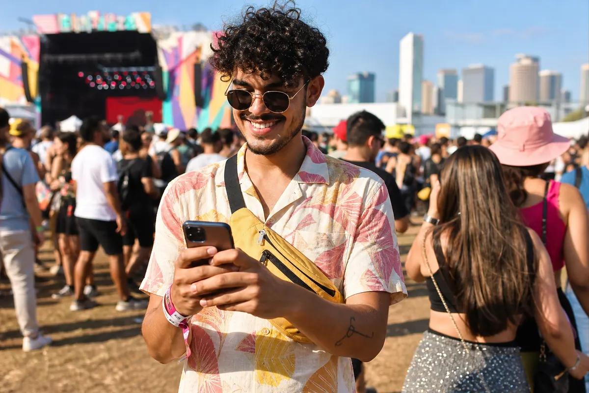 Smiling festival-goer holding his smartphone in the crowd, colorful stage and skyline behind