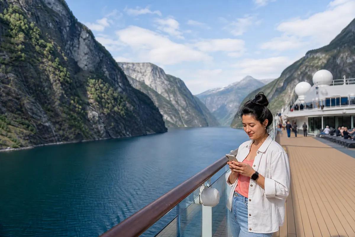 Female passenger checking her phone at the railing of a ship sailing through a Norwegian fjord