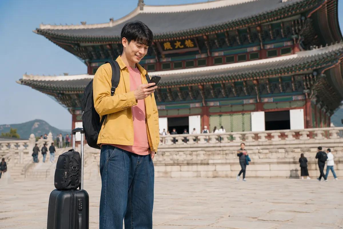 Smiling young traveler on his phone in front of a traditional Korean palace, suitcase at his feet