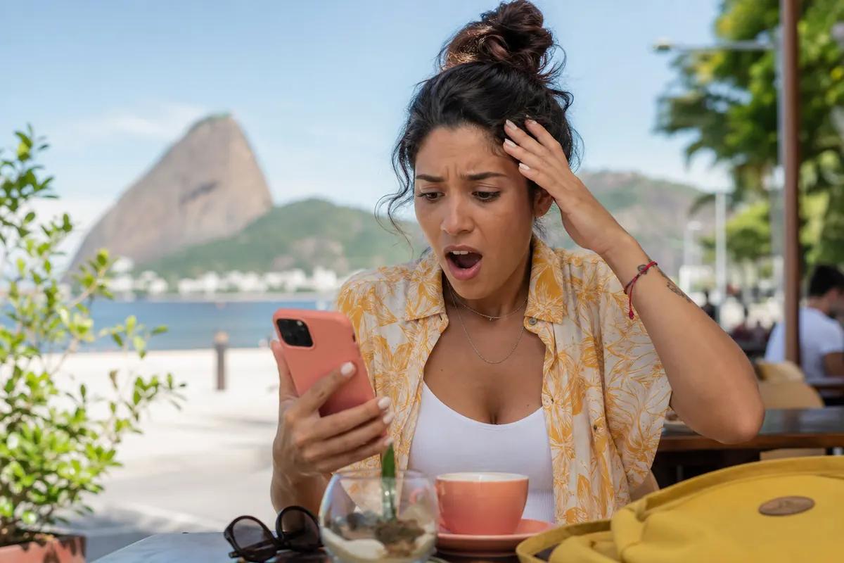 Stunned traveler staring at her phone on a cafe terrace with Rio's Sugarloaf Mountain behind