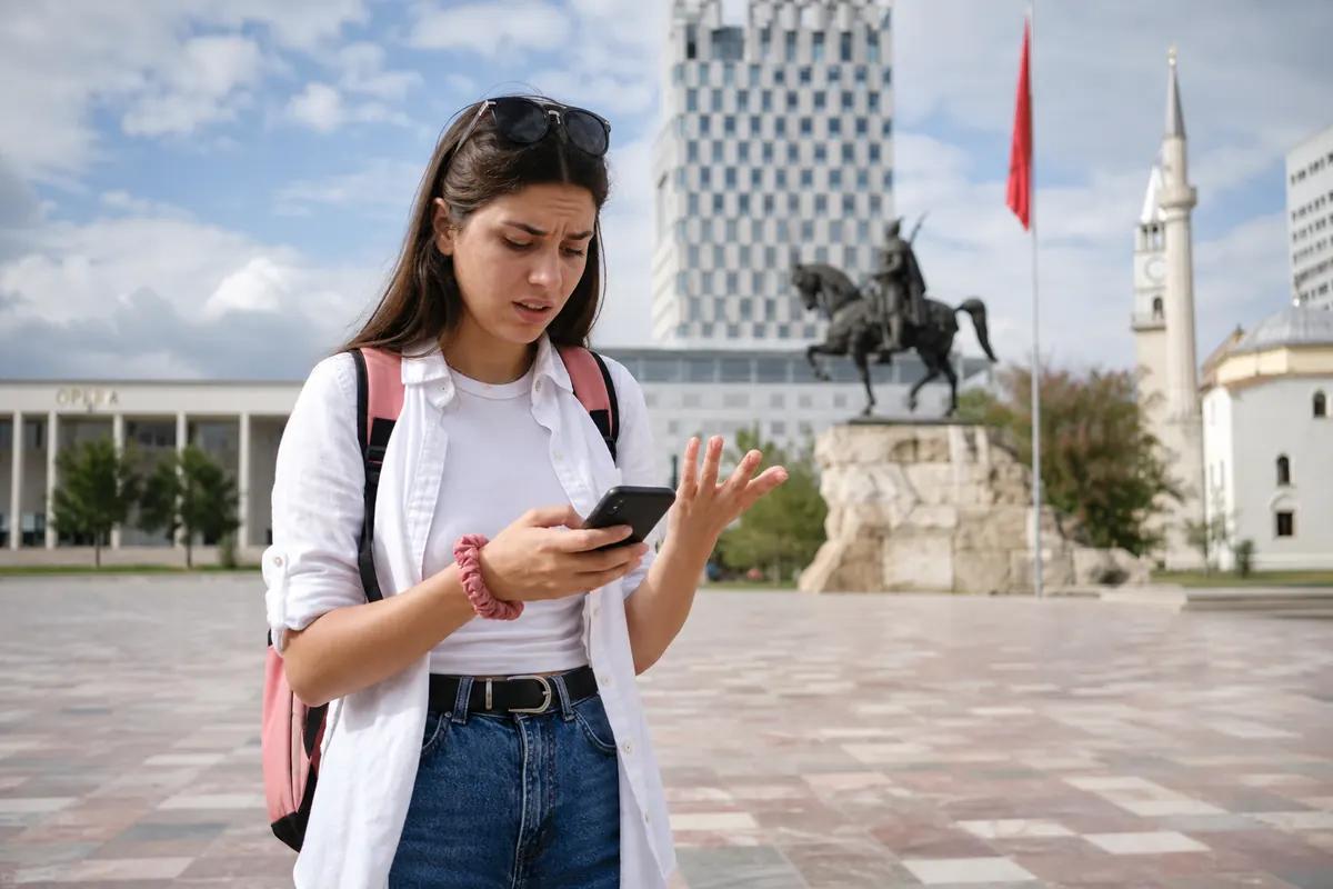 Confused female traveler looking at her phone on a Tirana square with an equestrian statue behind